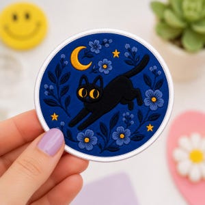 May include: A circular embroidered patch featuring a black cat with large yellow eyes leaping across a deep blue background. The design includes a yellow crescent moon, small yellow stars, and blue floral sprigs with yellow centers.