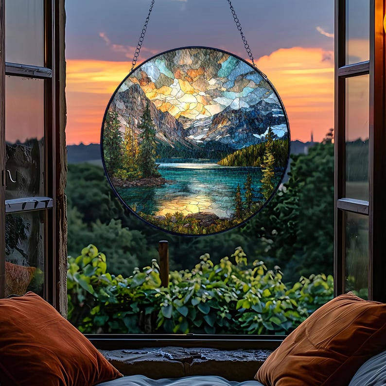 May include: A round stained glass window depicting a mountain lake scene, hanging in front of an open window. The glass art features a colorful landscape with mountains, trees, and water. The window is framed by a black metal border and chain.