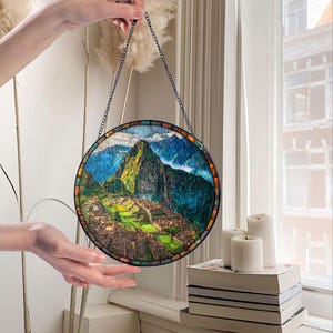 May include: Round stained glass panel depicting the Machu Picchu mountain landscape. The artwork features a detailed view of the ancient city with green and brown hues, framed by a mosaic border and a chain for hanging.