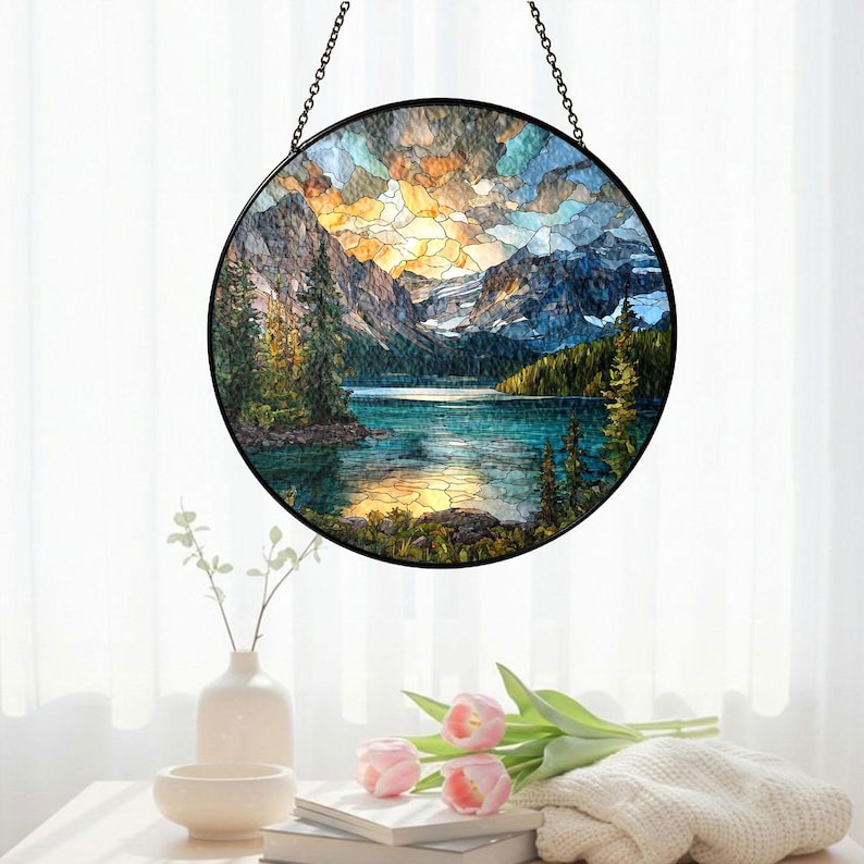 May include: Round stained glass window art depicting a mountain lake scene with a sunset. The artwork features a black metal frame and chain for hanging. The landscape includes mountains, trees, and a lake with a colorful sky. The window art is displayed in front of a white curtain.
