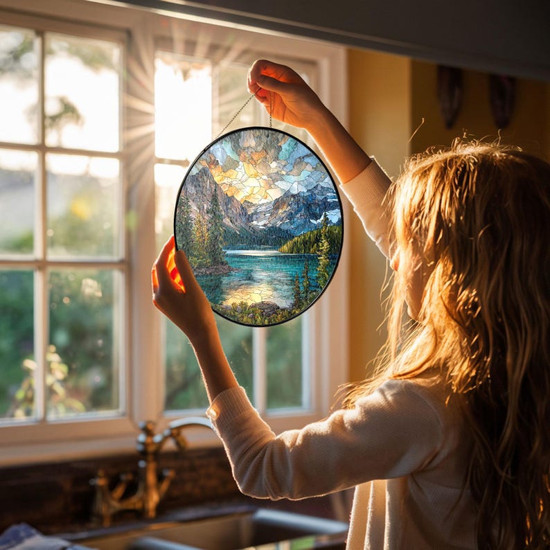 May include: A round stained glass panel depicting a mountain lake scene with a black metal frame. The artwork features a lake, mountains, and trees, with a colorful sky. The panel is held up in front of a window.