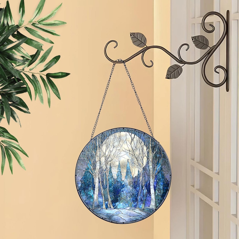 Op de afbeelding: Een rond gebrandschilderd raam met een winterbossc&egrave;ne met een volle maan. Het raam is zwart omlijst en hangt aan een decoratieve metalen ketting. Het raam is opgehangen aan een decoratieve metalen beugel met bladeren.