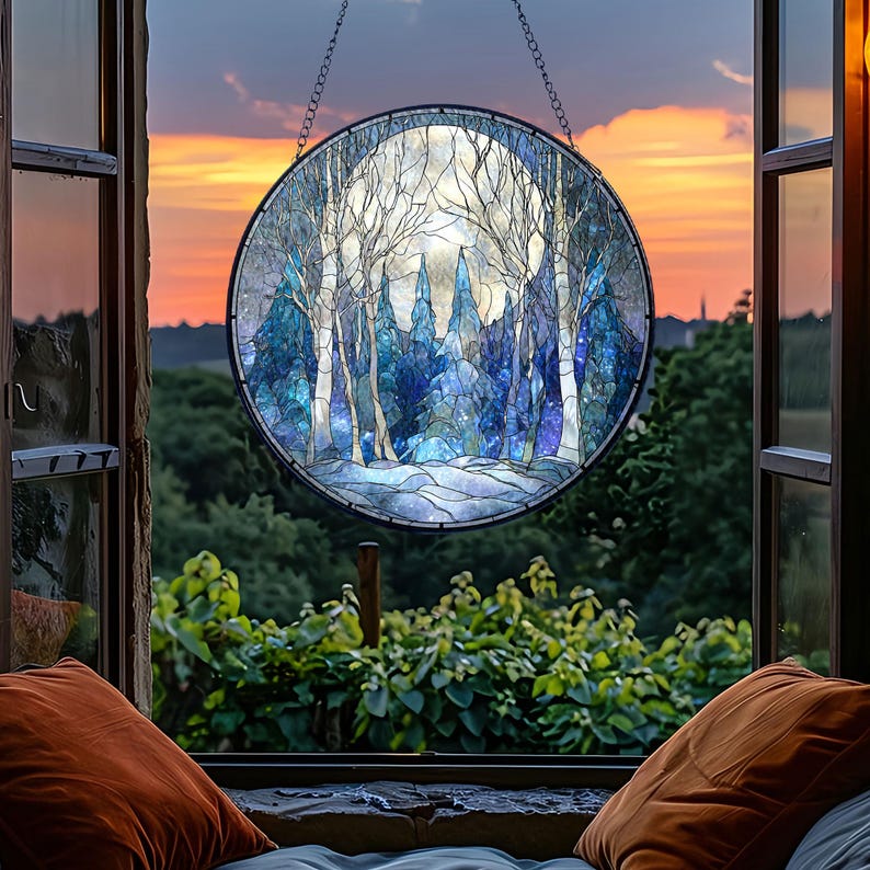Op de afbeelding: Een rond gebrandschilderd glazen raam dat een winterbossc&egrave;ne met een volle maan afbeeldt. Het kunstwerk heeft tinten blauw en wit, met kale bomen en een besneeuwde grond. Het raam hangt voor een open raam met uitzicht op de zonsondergang.