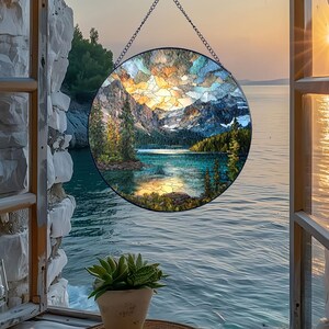 May include: A round stained glass panel depicting a mountain lake scene. The artwork features a vibrant sunset with orange, yellow, and blue hues. The panel is framed in black and suspended by a chain, showcasing a serene landscape.