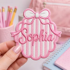 May include: A pink and white striped embroidered patch featuring a scalloped edge and a white bow at the top. The name Sophia is stitched in pink cursive script across the center of the patch. The design has a raised texture and pink border.