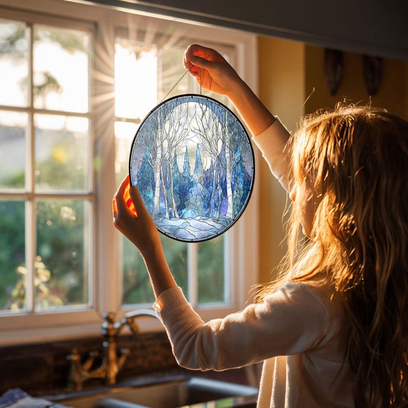 Op de afbeelding: Ronde gebrandschilderde glazen kunst met een winterbossc&egrave;ne met witte berken en een besneeuwde grond. Het kunstwerk heeft een zwart metalen frame en wordt opgehangen met een ketting. Het kleurenpalet omvat tinten blauw, wit en grijs.