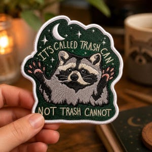 May include: An embroidered patch featuring a gray and black raccoon with raised paws against a dark green background. The design includes a white crescent moon, stars, and the text IT'S CALLED TRASH CAN NOT TRASH CANNOT in white lettering.