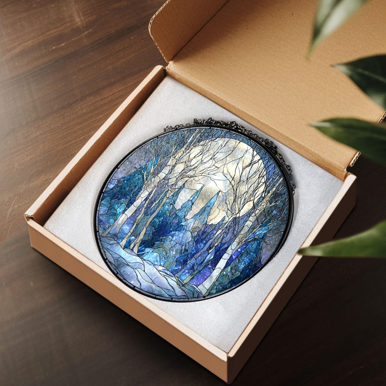 Op de afbeelding: Een rond gebrandschilderd glazen kunstwerk dat een maanverlicht bos toont met hoge, kale bomen en een heldere maan. Het kunstwerk is gevat in een zwart metalen frame en wordt gepresenteerd in een kartonnen doos met witte vulling.