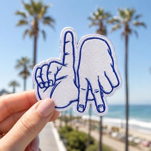 May include: An embroidered patch featuring a white hand gesture forming the letters L and A, outlined in bold blue thread. The design depicts the fingers positioned to create the iconic Los Angeles sign, with a clean white background and blue border.