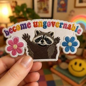 May include: An embroidered patch featuring a brown and black raccoon with raised paws, flanked by a pink flower on the left and a blue flower on the right. The text "become ungovernable" is stitched in multicolored letters above the raccoon.