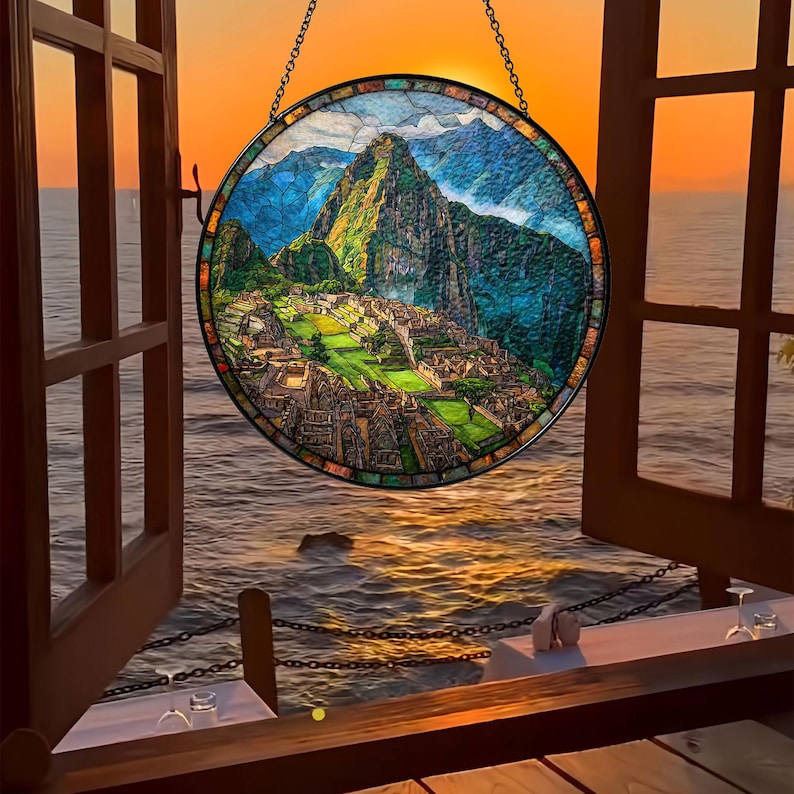 May include: A round stained glass window depicting Machu Picchu, hanging in front of an open window overlooking the ocean at sunset. The window features a detailed mosaic of the ancient city, mountains, and sky. The window is framed by a black metal chain.