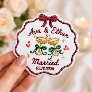 May include: A circular embroidered patch with a scalloped burgundy border, featuring a maroon bow at the top. It displays the text Ava & Ethan, Married, and 09.18.2026, with two gold champagne glasses, green ribbons, and small red hearts.