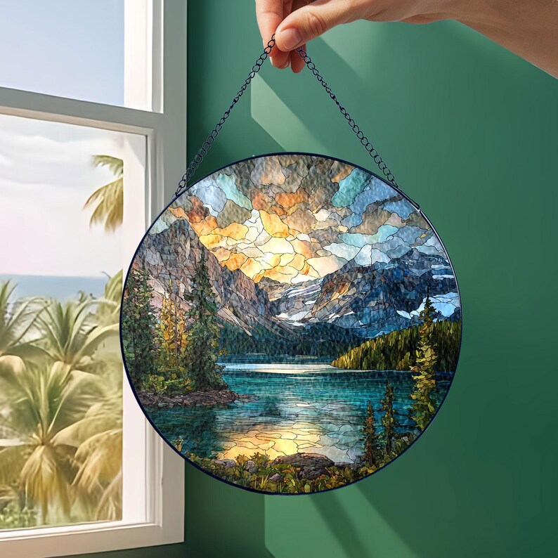 May include: Round stained glass panel depicting a mountain lake scene with a sunset. The artwork features a blue, orange, and yellow sky, with mountains and trees surrounding the lake. The panel is held by a chain.