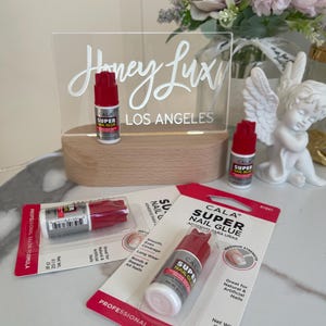 Puede incluir: Tres botellas de pegamento transparente para uñas con etiquetas rojas y blancas. Las etiquetas dicen "CALA SUPER NAIL GLUE" y "PROFESSIONAL SALON FORMULA". Las botellas están sobre un fondo blanco y rojo. Las botellas son de 0,1 oz (3 g).