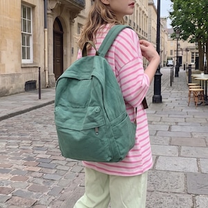 May include: A sage green canvas backpack with a large main compartment, a front zipped pocket, and two side pockets. The backpack features adjustable shoulder straps and a small top handle, worn over a pink and white horizontal striped t-shirt.