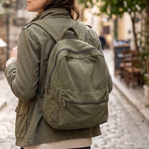 Könnte beinhalten: Ein olivgrüner Rucksack aus Segeltuch mit einem großen Hauptfach und einer kleineren Vordertasche, die beide mit dunkelbraunen Reißverschlüssen versehen sind. Der Rucksack verfügt über verstellbare Schultergurte und einen Tragegriff aus strukturiertem Stoff.