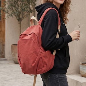 Könnte beinhalten: Ein terrakottafarbener Rucksack aus Segeltuch mit einem großen Hauptfach, einer Reißverschlusstasche auf der Vorderseite und Seitentaschen. Der Rucksack verfügt über hellbeige, verstellbare Schultergurte und einen Tragegriff oben.
