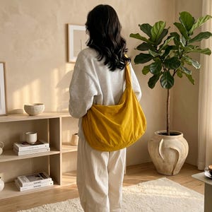 May include: A mustard yellow shoulder bag made of soft fabric. The bag is worn over the shoulder. The person is wearing a light gray sweatshirt and white pants. The background includes a plant in a large pot and a wooden shelf.