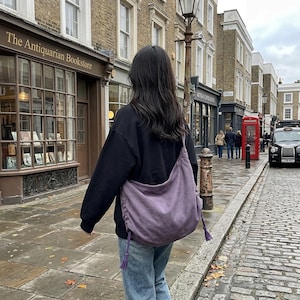 May include: A slouchy, purple hobo-style shoulder bag with a drawstring closure and tassel accents, worn over a black long-sleeve sweatshirt and blue denim jeans. The bag features a soft, unstructured fabric construction with visible side ties.