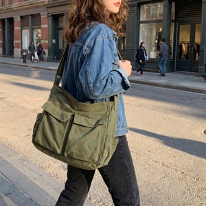 May include: An olive green canvas messenger bag with multiple front pockets and a long shoulder strap. The bag is worn over the shoulder. The bag is made of a durable material and is suitable for carrying books, laptops, or other essentials.