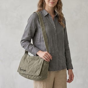 May include: An olive green canvas messenger bag with a fold-over flap and an adjustable shoulder strap. The bag features the name Treya embroidered in black script on the lower left corner of the front flap, providing a functional storage accessory.