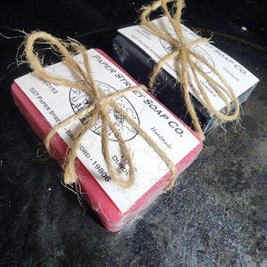 May include: Two rectangular bars of soap, one pink and one dark gray, wrapped in clear plastic. Each bar features a white paper label printed with the text Paper Street Soap Co., 537 Paper Street, and Tyler Durden, secured with brown twine.