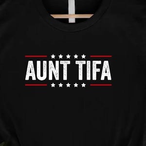 May include: Black t-shirt with the text "AUNT TIFA" in white, framed by red lines and stars. The shirt is hanging on a wooden hanger. The design is centered on the front of the shirt.