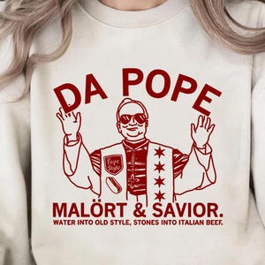 May include: A cream-coloured sweatshirt featuring a red graphic of a man wearing sunglasses and the text "DA POPE". The words "MALÖRT & SAVIOR." and "WATER INTO OLD STYLE, STONES INTO ITALIAN BEEF." are printed below.