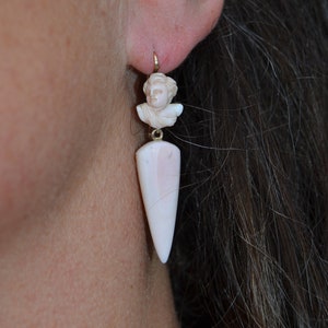 Puede incluir: Un par de pendientes de concha blanca con un acento dorado. Los pendientes presentan una pequeña figura tallada de la cabeza de una persona en la parte superior y una concha larga, puntiaguda, en forma de lágrima en la parte inferior.