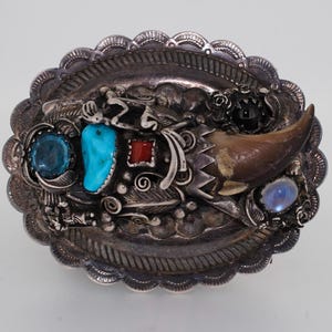 May include: Oval silver belt buckle with intricate details. Features a turquoise stone, a blue stone, a red stone, and a brown claw. The buckle has a scalloped edge and decorative silverwork.