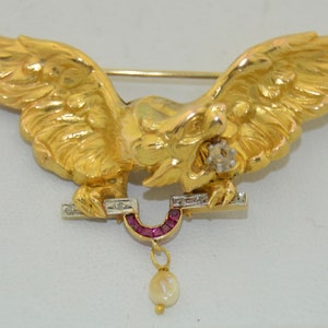 Puede incluir: Un broche dorado con un grifo alado detallado con un acento de rubí y diamante. El grifo sostiene un colgante en forma de herradura con una gota de perla.