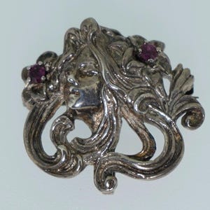 May include: A silver-toned brooch featuring a woman's face with flowing hair and floral accents. Two small, round, red gemstones are set within the floral details. The brooch has an Art Nouveau style.