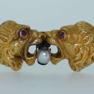May include: A gold-toned brooch featuring two lion heads facing each other, with ruby eyes and open mouths. A pearl is held between their jaws. The brooch has intricate detailing and a vintage aesthetic.