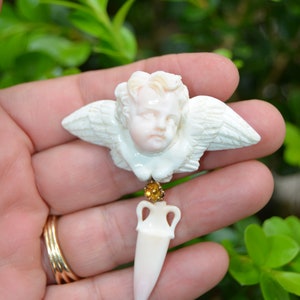 Puede incluir: Un querubín de porcelana blanca con alas y un delicado colgante de porcelana blanca puntiagudo que cuelga de una cuenta dorada. El querubín sostiene un pequeño jarrón de porcelana blanca.