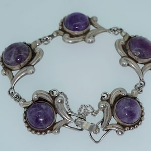 May include: Silver bracelet with five oval, deep purple gemstones. The bracelet has an ornate, floral design with a chain and clasp closure. The gemstones are set in decorative bezels. The bracelet is approximately 18 cm in length.