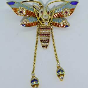 May include: A gold-toned butterfly brooch with blue, orange, and red enamel wings. The brooch features ruby and diamond accents, with dangling chain details ending in blue and gold enamel teardrops.