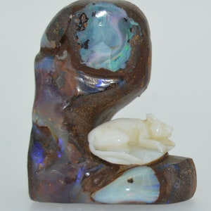 Puede incluir: Una escultura de opalo tallada de un ratón sentado en una hoja. El opalo es de color marrón oscuro con destellos iridiscentes azules, verdes y morados. El ratón está tallado en opalo blanco.