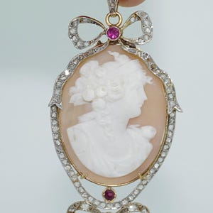 May include: A cameo pendant with a white cameo of a woman's profile set in a gold frame with a pink background. The pendant is adorned with a bow at the top and bottom, each with a pink stone in the center and surrounded by small diamonds.