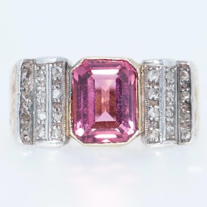 May include: A pink gemstone ring with a square cut stone set in a gold band. The band is surrounded by a row of small diamonds.