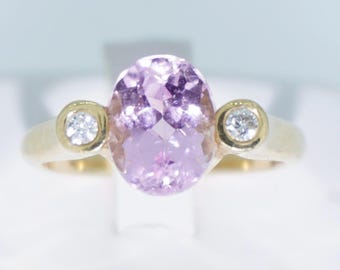 Può includere: Un anello d'oro con una grande pietra ovale viola chiaro e due pietre preziose rotonde e trasparenti più piccole. Il design dell'anello è classico ed elegante. Le pietre sono incastonate in una fascia d'oro.