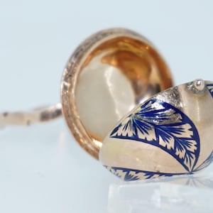 Puede incluir: Un anillo de oro con una parte superior en forma de huevo de esmalte azul y blanco con bisagras. El huevo está abierto, revelando el interior dorado.