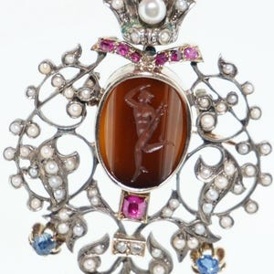 Puede incluir: Un colgante de plata con un camafeo que presenta una figura tallada, rodeado de perlas y rubíes. El colgante tiene la forma de un diseño floral estilizado.