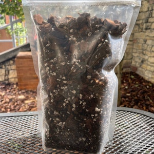 May include: A clear plastic bag filled with a dark brown potting mix. The mix contains pieces of bark, dark soil, and small white particles. The bag is sealed and sits on a metal table.