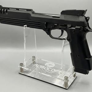 May include: A black handgun with a ribbed slide and a prominent barrel, displayed on a clear acrylic stand. The stand features the text "ROBOCOP" and a logo. The gun is a replica or prop.