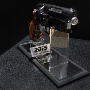 May include: A black and orange handgun on a clear acrylic stand with a mirrored base. The stand has a silver label that reads "2019 DETECTIVE SPECIAL". The gun is displayed against a dark background.