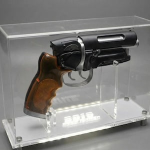 May include: A black and silver handgun with a brown grip is displayed inside a clear acrylic case. The gun is mounted on a white base with the text "2012" visible. The case is rectangular and has a clear top.