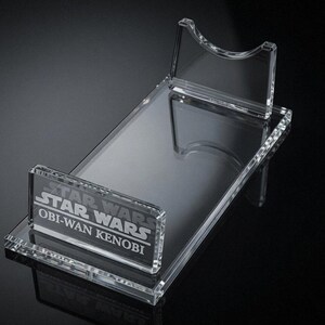 May include: Clear acrylic display stand for a Star Wars Obi-Wan Kenobi collectible. The stand features a rectangular base with a raised platform and a back support. The Star Wars logo and character name are etched on the front.