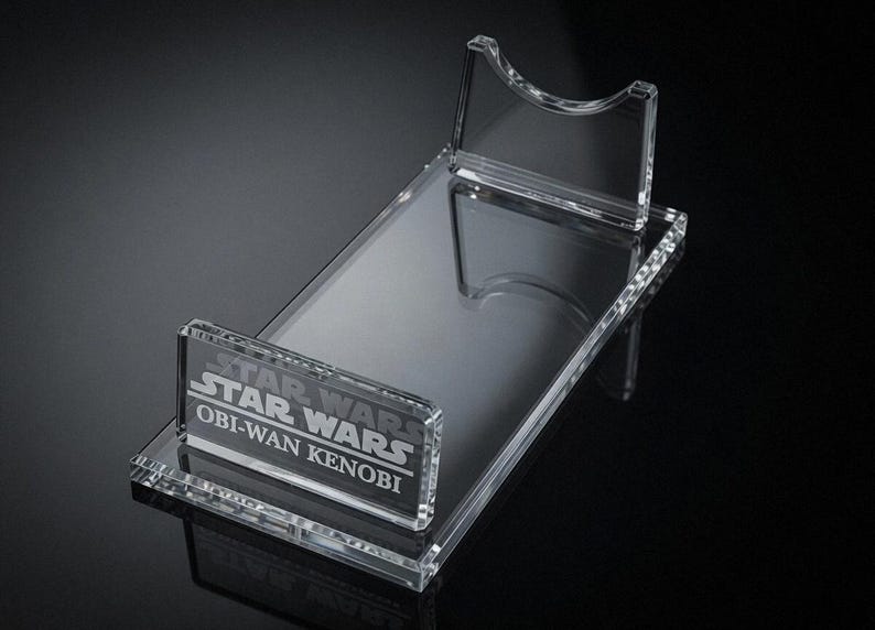 May include: Clear acrylic display stand for a Star Wars Obi-Wan Kenobi collectible. The stand features a rectangular base with a raised platform and a back support. The Star Wars logo and character name are etched on the front.