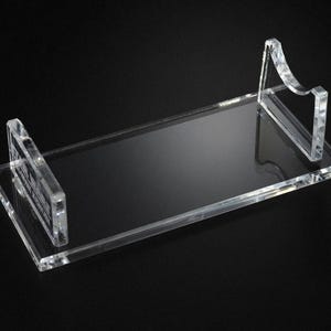 May include: A clear acrylic display stand with a rectangular base and two upright supports. The supports have a curved design. The stand is designed to hold an item and is set against a black background.
