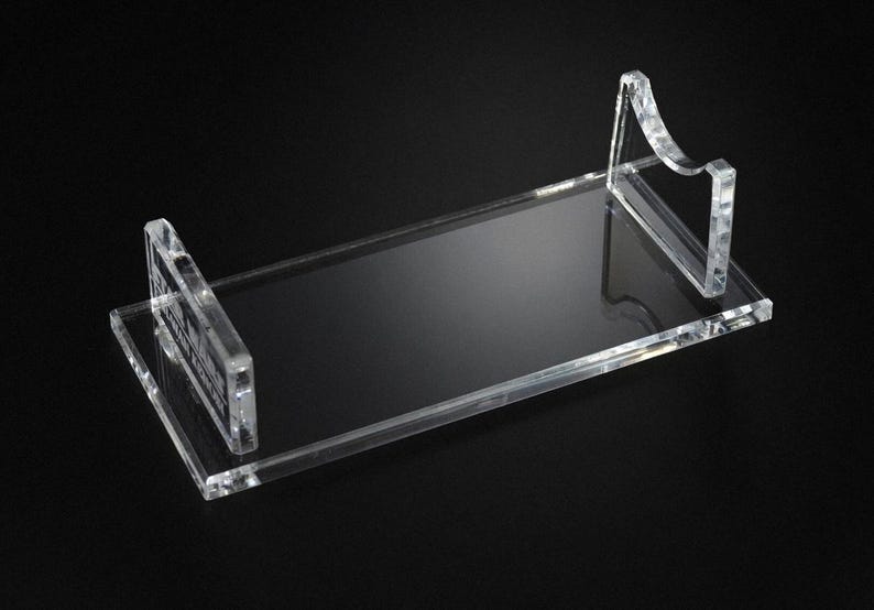 May include: A clear acrylic display stand with a rectangular base and two upright supports. The supports have a curved design. The stand is designed to hold an item and is set against a black background.